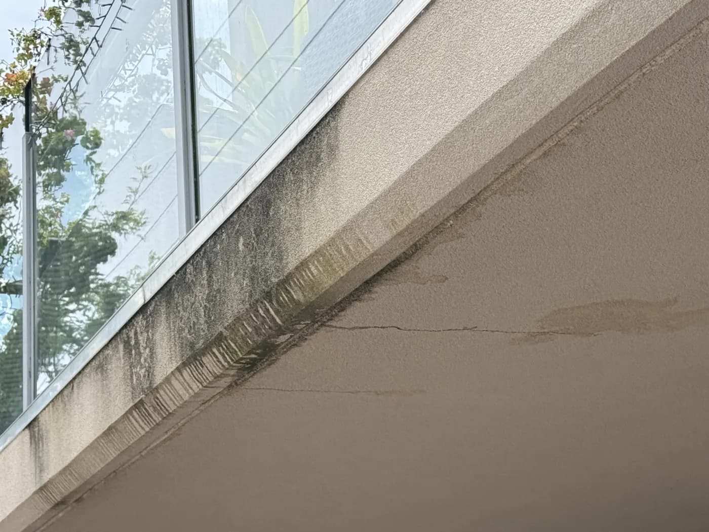 Rendered facade below a glazing line, dark with algae where rain sheets down it, and a crack running through the render
