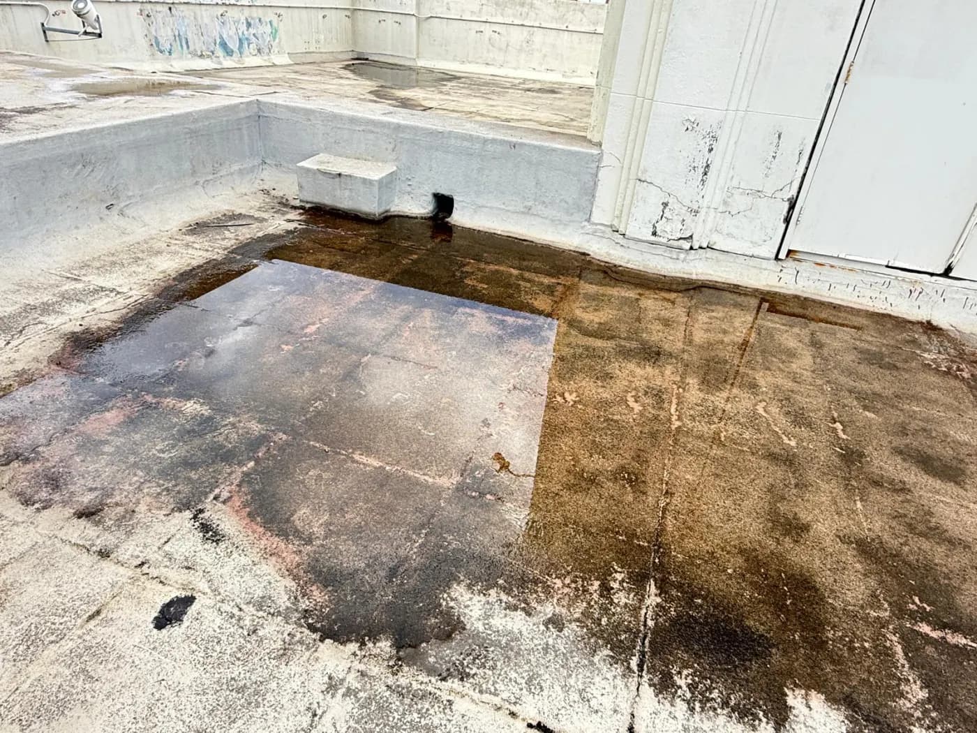 Bare concrete deck holding a wide pool of standing water against an upstand, heavily stained from doing so for a long time
